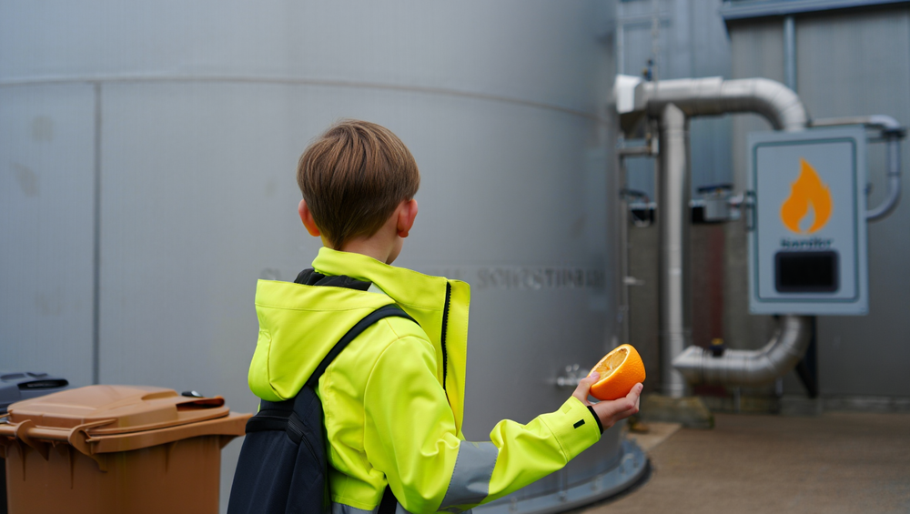 Bambino visto di spalle, con giubbetto ad alta visibilità, tiene in mano una buccia d’arancia davanti a un biodigestore industriale.
