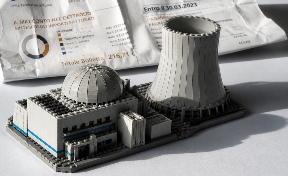 Modello LEGO iperrealistico di una centrale nucleare, con cupola del reattore e torre di raffreddamento, illuminato da luce direzionale su fondo bianco.