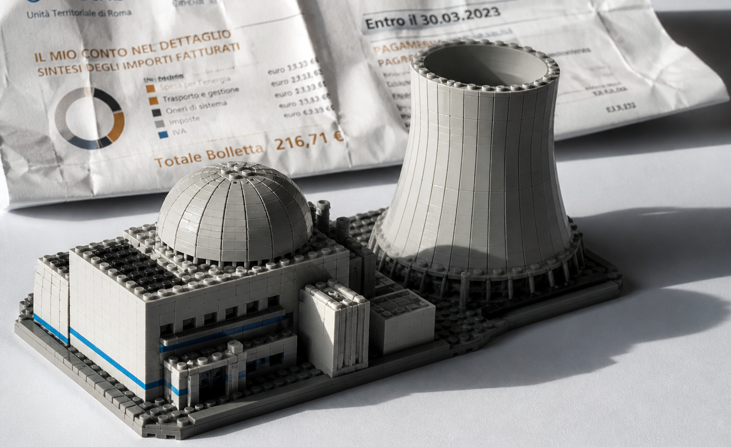 Modello LEGO iperrealistico di una centrale nucleare, con cupola del reattore e torre di raffreddamento, illuminato da luce direzionale su fondo bianco.