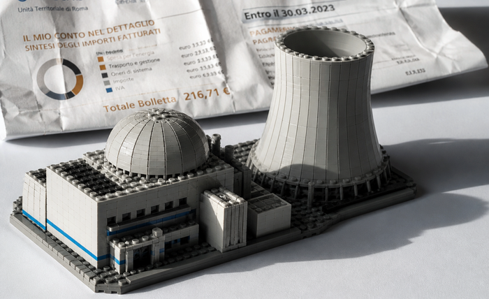 Modello LEGO iperrealistico di una centrale nucleare, con cupola del reattore e torre di raffreddamento, illuminato da luce direzionale su fondo bianco.